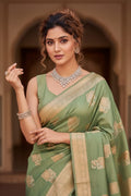 Woman in a green saree with gold patterns, wearing jewelry, against an architectural background.
