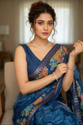 Woman in a blue saree with floral patterns sitting indoors.