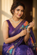 Woman in a purple saree with floral patterns, wearing jewelry, indoors.