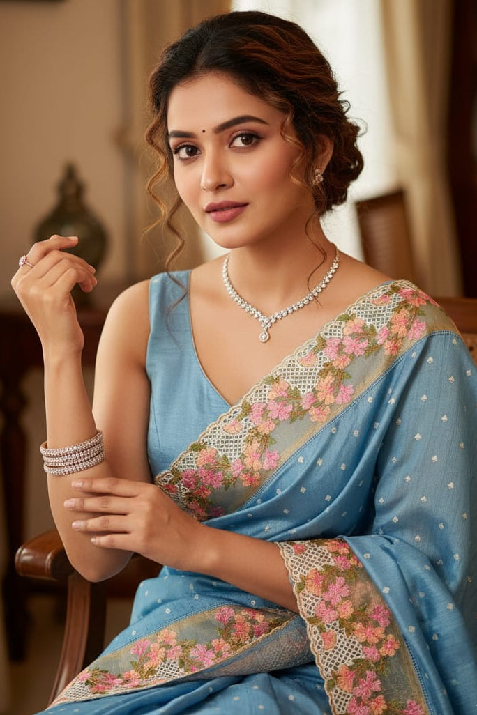 Woman in a blue saree with floral patterns sitting indoors.