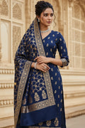 Woman wearing a blue traditional outfit with intricate patterns in front of an architectural background.