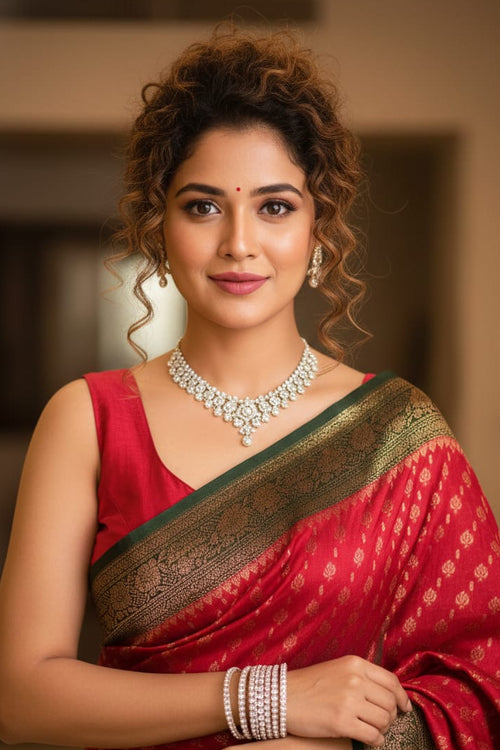 Woman in a red saree with gold border, wearing jewelry, against a blurred indoor background
