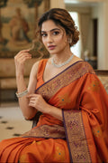 Woman in an orange saree with intricate patterns in a decorated room.